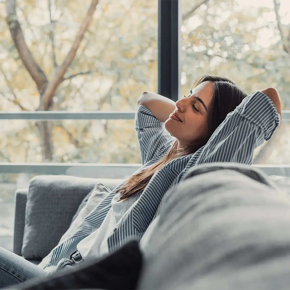 Vrouw die ontspant op een bank bij een groot raam &ndash; lichte interieur, akoestisch comfort en een gevoel van veiligheid in een modern huis.